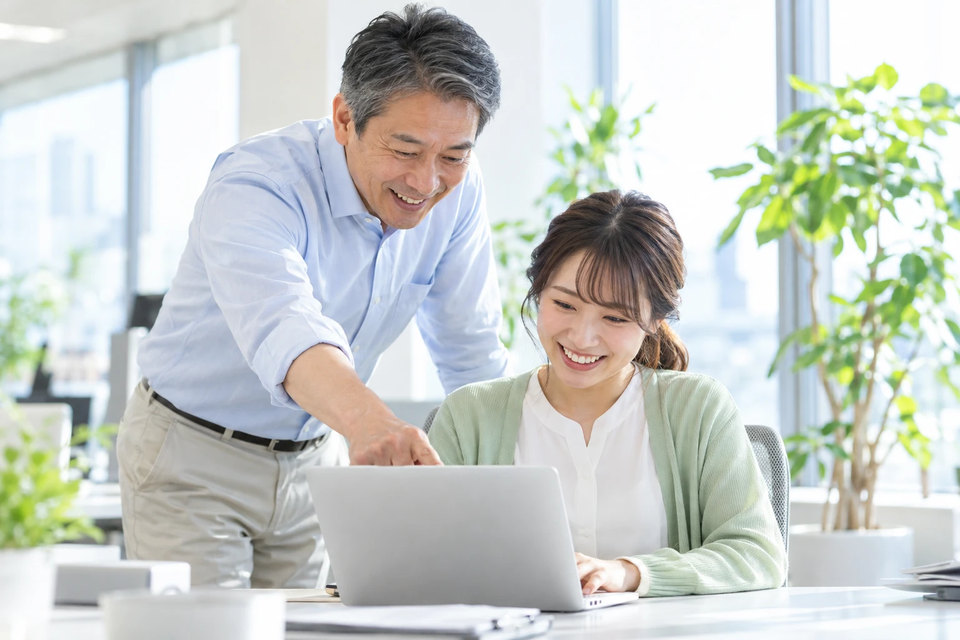 明るいオフィスの席で、営業担当のノートパソコンの画面を指差しながら笑顔で助言する営業部長