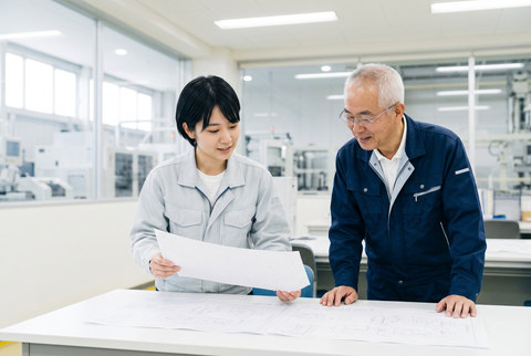 印刷した図面を手に取って広げる若手技術者と、机に手をついてのぞき込むベテラン技術者