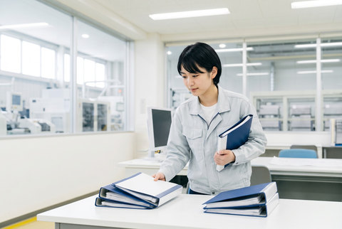 設計室の机で、資料のバインダーを2つの山に分けて整理する若手技術者