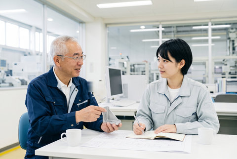打ち合わせの机で、金属の部品を手に説明するベテラン技術者と、ノートにメモを取る若手技術者