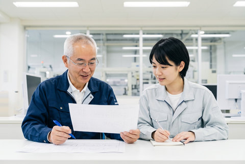 印刷した資料にペンを当てて確かめるベテラン技術者と、隣でノートに書き留める若手技術者