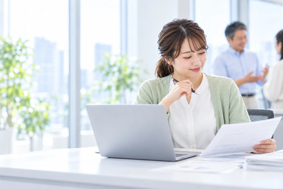明るいオフィスの席で、ノートパソコンの横に置いた提案書を見直す営業担当。奥の窓ぎわでは営業部長が同僚と話している