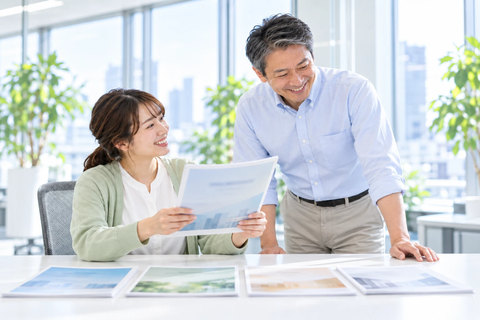 窓ぎわの席で、机に並べた提案書から1部を選んで部長に見せる営業担当と、笑顔でのぞき込む営業部長