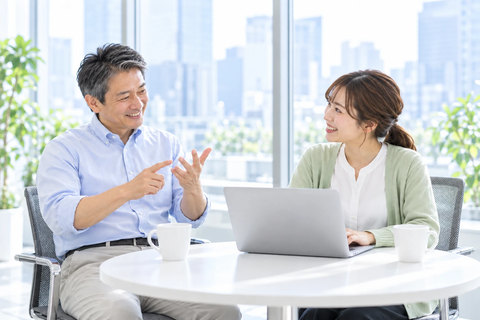 窓ぎわの丸テーブルで、指を折りながら話す営業部長と、ノートパソコンでメモを取る営業担当